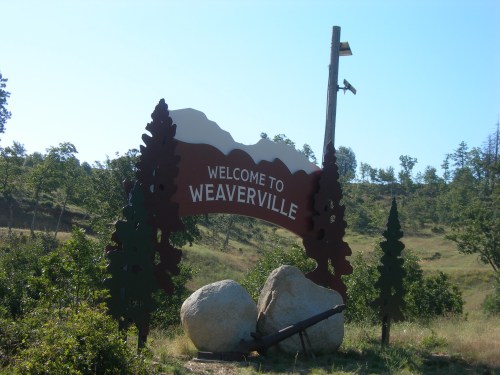 Welcome sign for Weaverville, featuring mountains and trees, set in a green landscape under a clear blue sky.