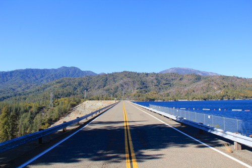 A straight road lined with guardrails, flanked by a lake and mountains under a clear blue sky.