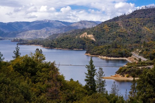 A serene lake surrounded by mountains and trees under a partly cloudy sky. A road runs along the water's edge.