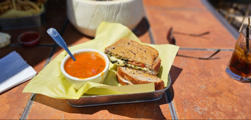 A grilled sandwich with herbs and tomato, served with a bowl of tomato soup on a sunny outdoor table.