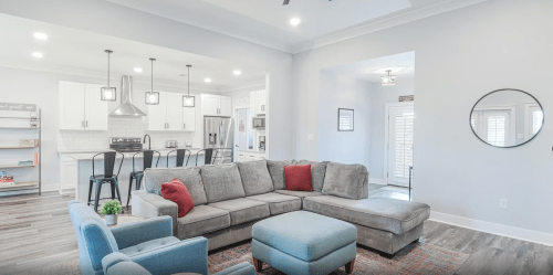 Modern living room with a gray sectional sofa, blue accent chair, and open kitchen in the background. Bright and airy space.