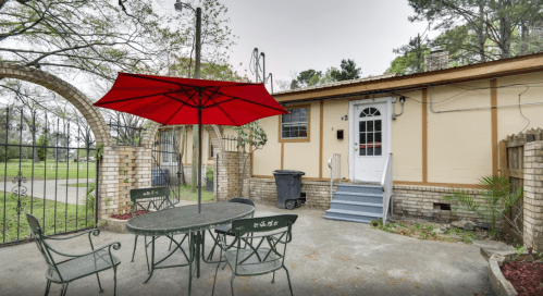 A patio with a red umbrella over a table and chairs, next to a brick wall and a gated entrance.