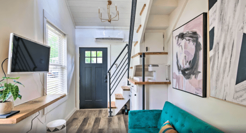 A cozy interior featuring a teal couch, wall art, a TV, and a staircase with wooden shelves in a bright room.