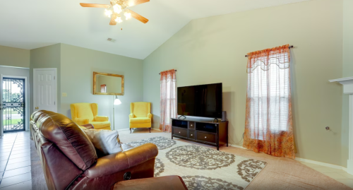 Cozy living room with yellow armchairs, a TV, and a decorative rug, featuring large windows and a ceiling fan.
