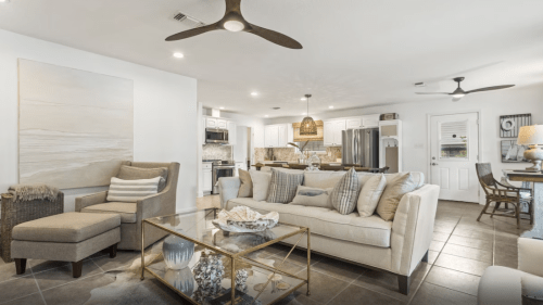 A modern living room with a beige sofa, glass coffee table, and open kitchen, featuring light decor and ceiling fans.