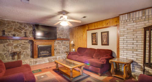 Cozy living room with stone fireplace, wooden accents, red sofas, and a coffee table under a ceiling fan.