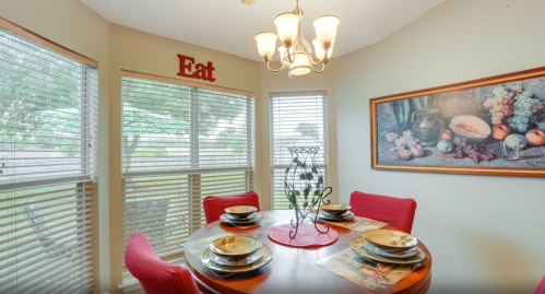 A cozy dining area with a round table set for four, featuring a chandelier and large windows with greenery outside.
