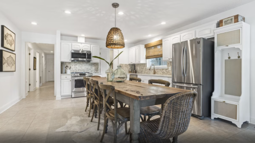 A modern kitchen with a wooden dining table, wicker chairs, and stainless steel appliances, featuring bright lighting.