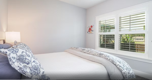 Cozy bedroom with a neatly made bed, light gray walls, and a window with white shutters letting in natural light.