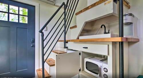 A small kitchen area with a microwave, sink, and stairs leading to an upper level, next to a dark blue door.