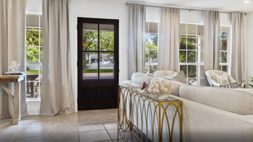 Bright living room with large windows, light curtains, a cozy sofa, and a decorative table near a front door.