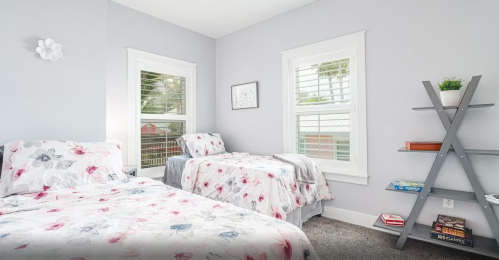 Bright bedroom with two beds, floral bedding, and a modern bookshelf against a light gray wall. Natural light from windows.