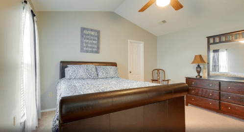 Cozy bedroom with a bed, dresser, chair, and a wall sign that reads "Happiness." Bright natural light from the window.