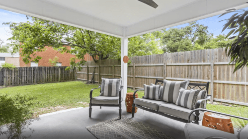 A cozy patio with a seating area, decorative pillows, and a view of a green backyard and wooden fence.