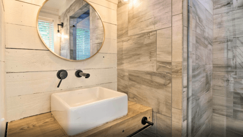 Modern bathroom featuring a round mirror, a white sink on a wooden countertop, and a tiled shower area.