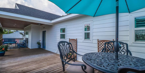 A patio with a round table, chairs, and a large umbrella, featuring wooden decking and white walls.