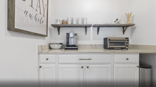 A modern kitchen corner with white cabinets, a coffee maker, toaster, and shelves holding glassware and decor.