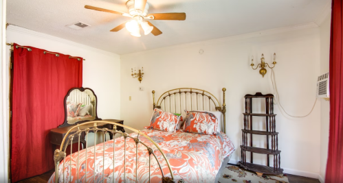 Cozy bedroom with a floral bedspread, red curtains, a mirror, and a wooden shelf against a white wall.
