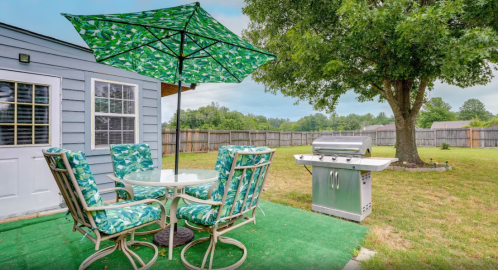 A backyard patio with a glass table, four chairs under a green umbrella, and a grill beside a large tree.