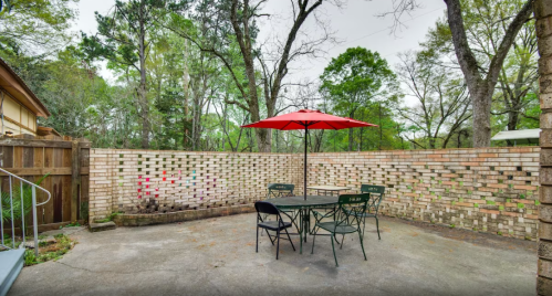 A patio with a red umbrella over a table and chairs, surrounded by a brick wall and greenery in the background.