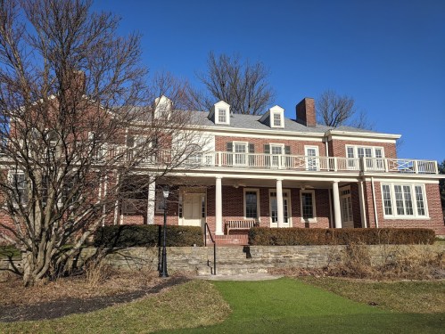 A large brick house with a white balcony, surrounded by trees and a green lawn under a clear blue sky.
