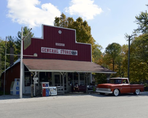 A red general store with a vintage truck parked outside, surrounded by trees and a clear blue sky.