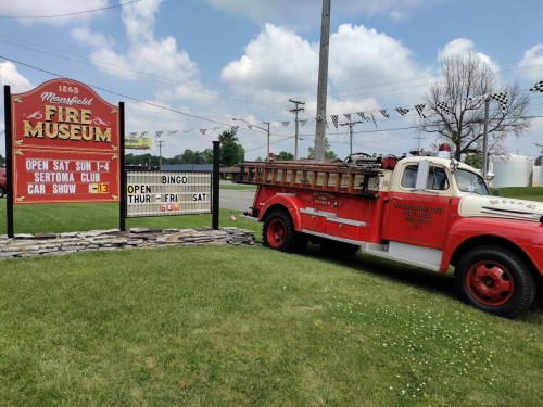 A vintage fire truck parked beside the Marshall Fire Museum sign, featuring open hours and event details.