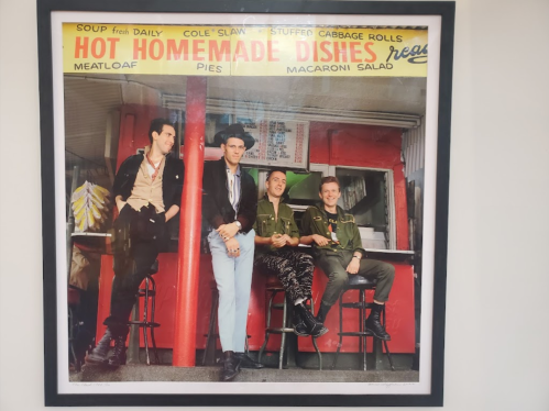 Four men pose in front of a colorful diner sign advertising homemade dishes, including meatloaf and pies.
