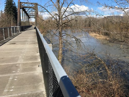 A walkway on a bridge overlooking a river, surrounded by trees and a clear blue sky with scattered clouds.