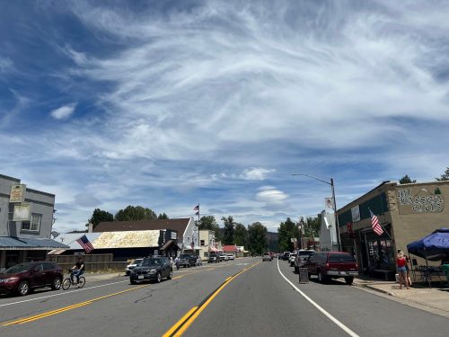 A small town street lined with shops, cars, and American flags under a blue sky with wispy clouds.