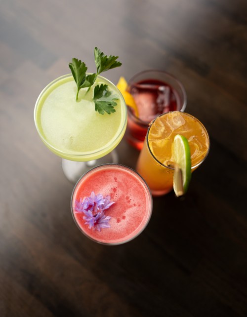 Four colorful cocktails on a wooden table, featuring garnishes like herbs and flowers.