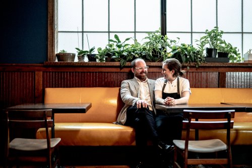 A man and woman sit together on a yellow bench in a cozy café, surrounded by plants and natural light.