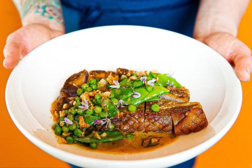 A person holds a white bowl filled with sliced meat, green peas, and garnished with herbs against an orange background.