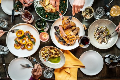 A table filled with various dishes, including roasted meats, vegetables, and drinks, with hands reaching for food.
