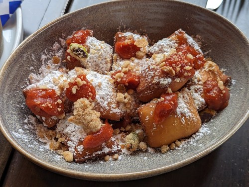 A bowl of sweet dumplings topped with powdered sugar, crushed nuts, and a red sauce.