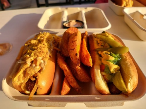 Two hot dogs topped with mustard and pickles, served with sweet potato fries in a takeout container.