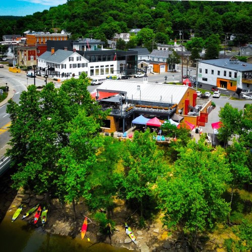 Aerial view of a riverside town with colorful buildings, trees, and kayaks along the water's edge.