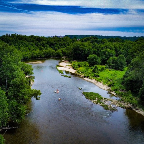 Aerial view of a winding river surrounded by lush green trees and a sandy bank, with kayakers paddling along.