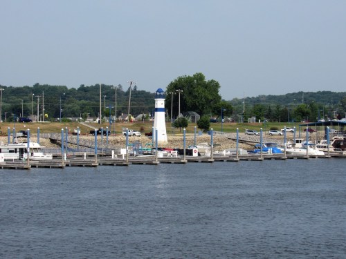 A marina with boats docked, featuring a blue and white lighthouse in the background and green trees along the shore.