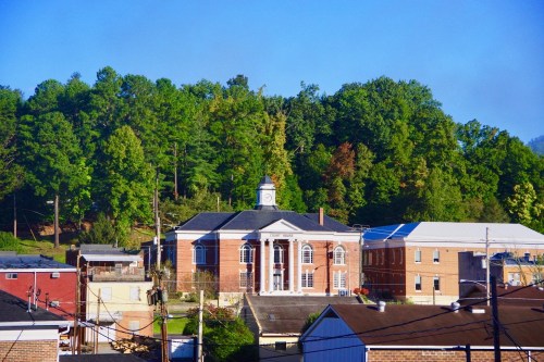 Historic brick building with a clock tower, surrounded by trees and other structures under a clear blue sky.