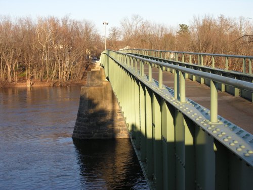 A green metal bridge spans a river, with trees lining the banks and a clear sky in the background.