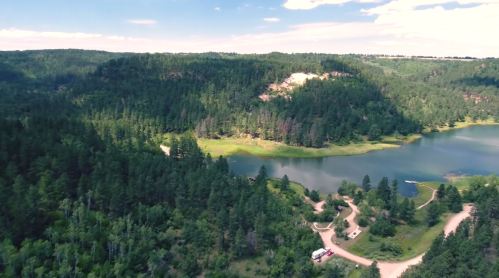 Aerial view of a lush green landscape with a lake, surrounded by hills and trees under a clear blue sky.