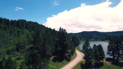 A scenic view of a winding dirt road beside a lake, surrounded by lush green trees and a blue sky with clouds.