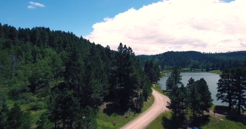 Aerial view of a winding dirt road beside a serene lake, surrounded by lush green trees and a blue sky with clouds.