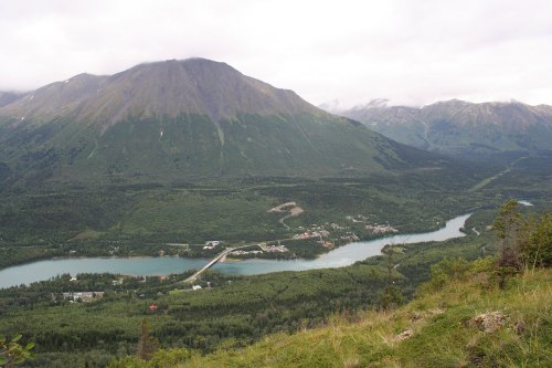 A scenic view of a river winding through lush green valleys, surrounded by mountains under a cloudy sky.