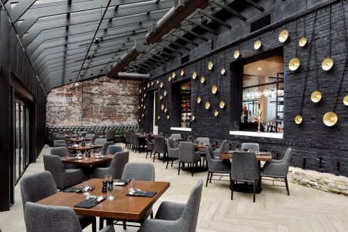 Modern restaurant interior with wooden tables, gray chairs, and decorative lighting against a brick wall.