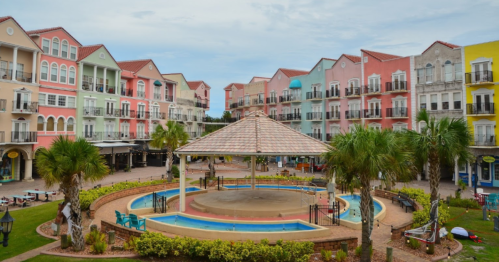 Colorful buildings surround a central gazebo and fountain in a vibrant plaza, with palm trees and seating areas.