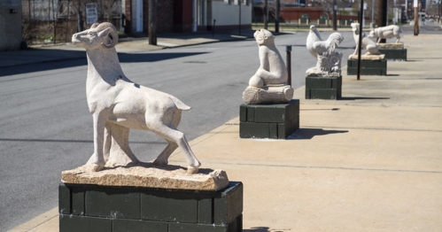 A row of stone sculptures depicting a goat, a seated figure, and a rooster along a sidewalk.