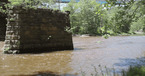 A stone pillar stands by a muddy river, surrounded by lush green trees and a clear blue sky.