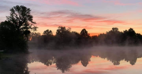 A serene lake at sunrise, with mist rising and trees reflecting on the calm water under a colorful sky.